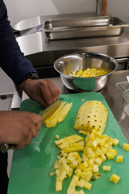 Auf einer grünen Schneideunterlage werden von Händen mit einem Messer Ananasstücke geschnitten. Eine Schüssel gefüllt mit gewürfelten Ananasstücken ist im Hintergrund zu sehen. Edelstahlflächen sind ebenfalls sichtbar.